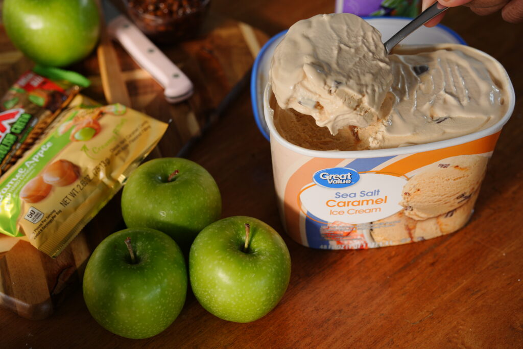 A spoon scooping creamy Great Value Sea Salt Caramel Ice Cream beside fresh green apples and caramel apple candy bars, ready to blend into a fall milkshake.