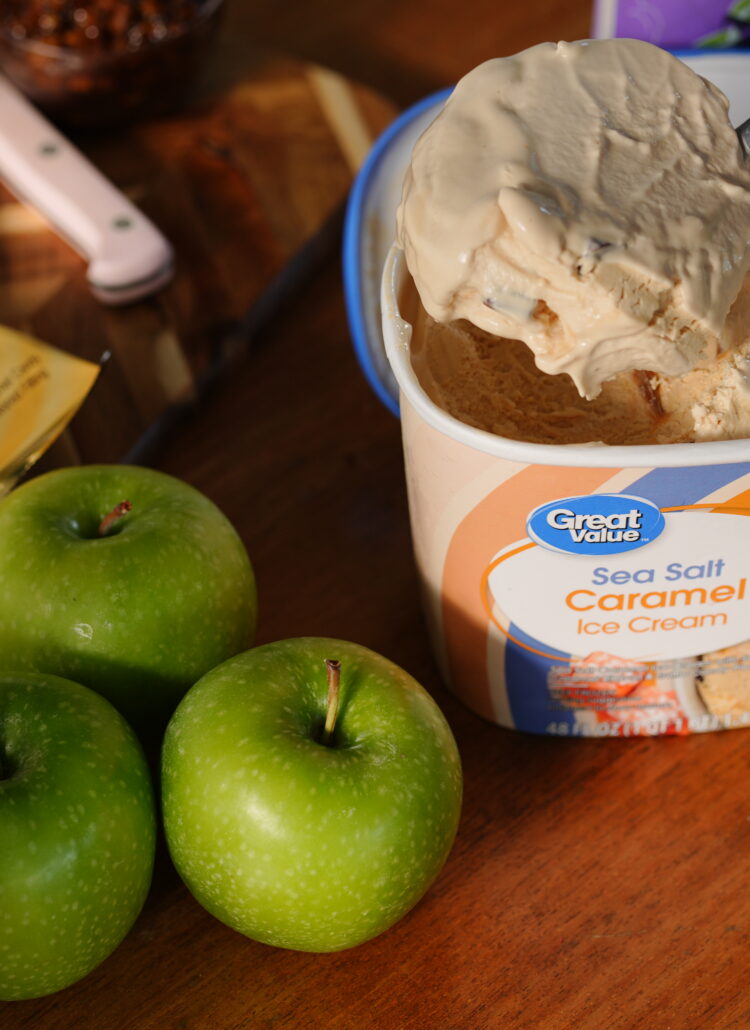 A spoon scooping creamy Great Value Sea Salt Caramel Ice Cream beside fresh green apples and caramel apple candy bars, ready to blend into a fall milkshake.