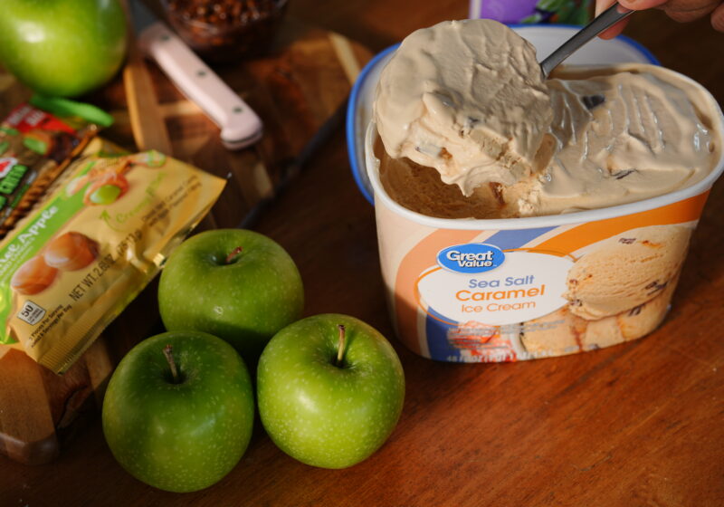 A spoon scooping creamy Great Value Sea Salt Caramel Ice Cream beside fresh green apples and caramel apple candy bars, ready to blend into a fall milkshake.