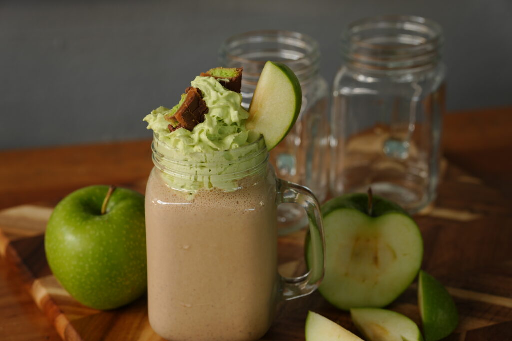 A mason jar glass filled with creamy caramel apple milkshake topped with caramel apple whipped cream, Twix pieces, and a slice of Granny Smith apple.