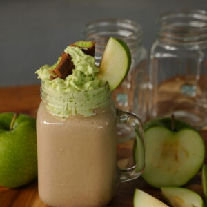 A mason jar glass filled with creamy caramel apple milkshake topped with caramel apple whipped cream, Twix pieces, and a slice of Granny Smith apple.