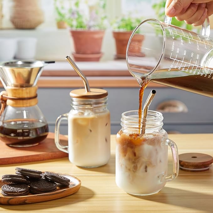 Pouring coffee into a DRASTAR iced coffee cup with milk and ice, featuring stainless steel straws, acacia lids, and a cozy kitchen setup with cookies on the counter.